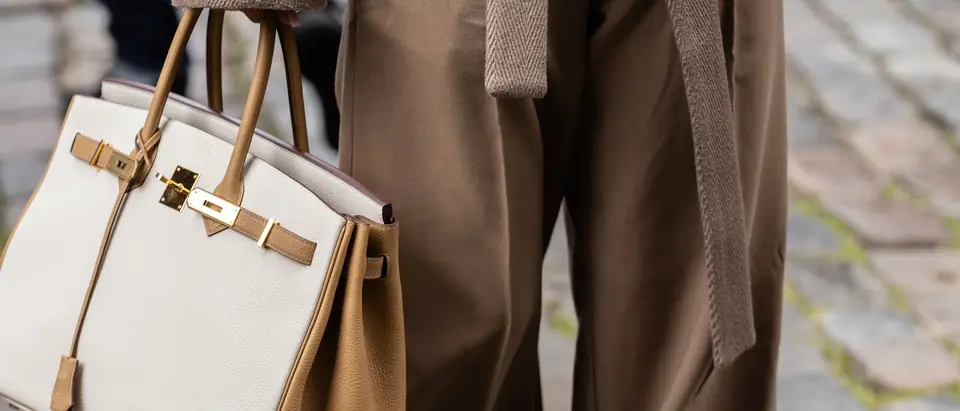 A woman in a camel coat carrying a white and gold Hermes Birkin on a cobbled street