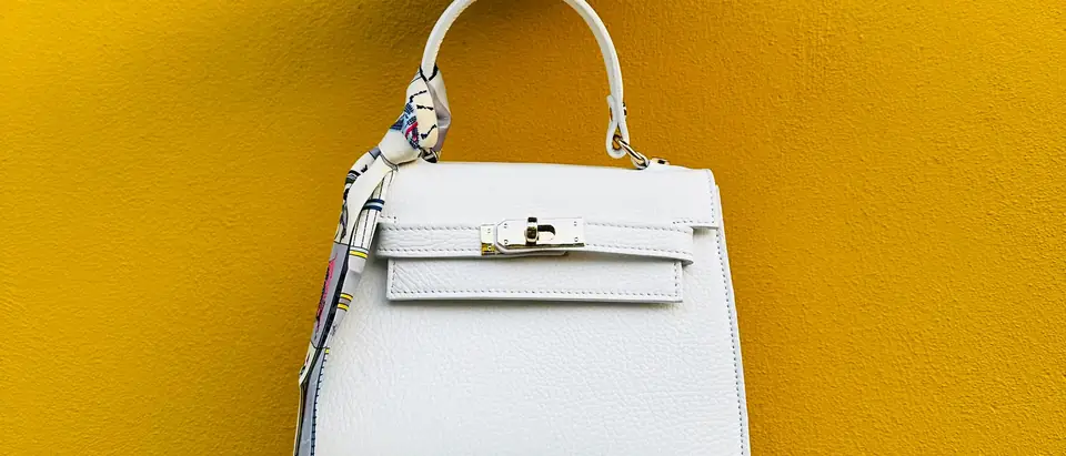 A white Hermes Kelly bag with a silk twilly held up against a bright yellow wall